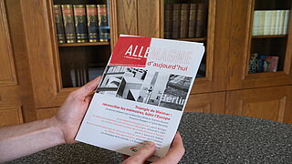 Zwei Hände halten Druckausgabe von Allemagne d’aujourd’hui, auf der man das Thema sieht. Im Hintergrund stehen Bücherregale.