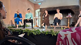 Podiumsdiskussion auf einer Bühne. Vier Menschen nehmen teil, eine Frau spricht ins Mikrofon.